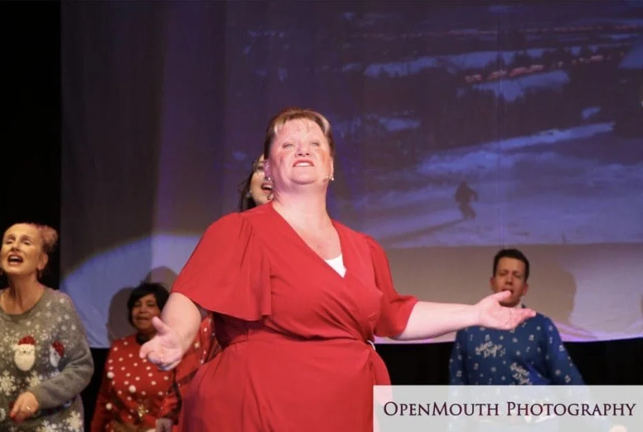 A woman in a red dress performing on stage with a joyful expression, surrounded by other people in holiday sweaters, with a snowy winter scene projected behind them.