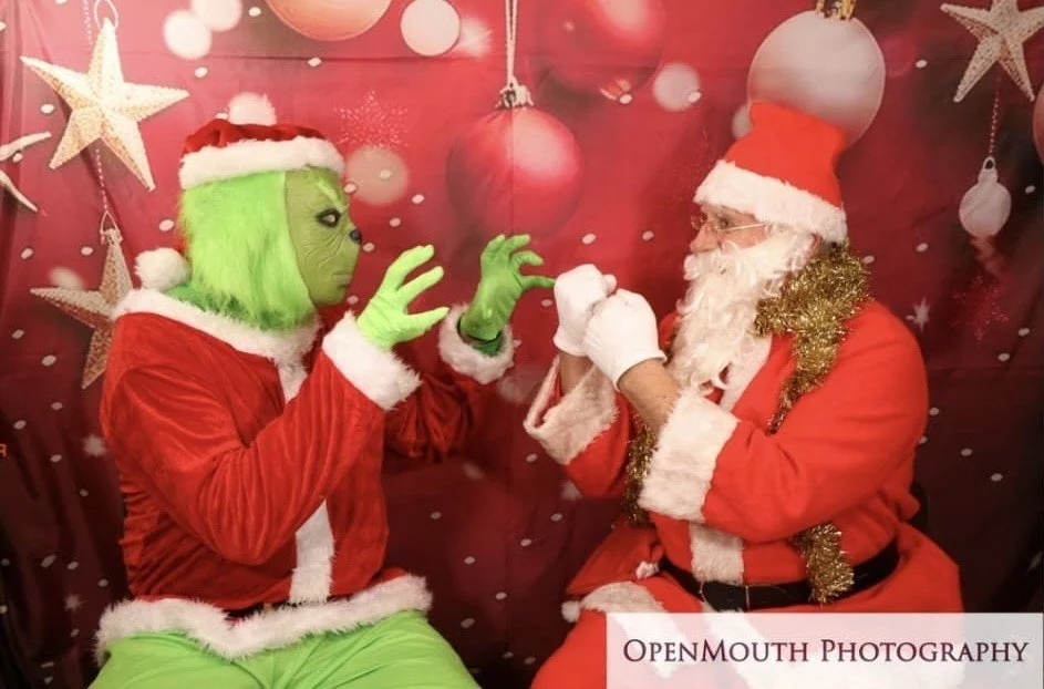 A person dressed as the Grinch in a red Santa suit with green face and hands talking to a man dressed as Santa Claus with glasses, a white beard, and a red suit with gold tinsel, in front of a red background decorated with white ornaments and stars.