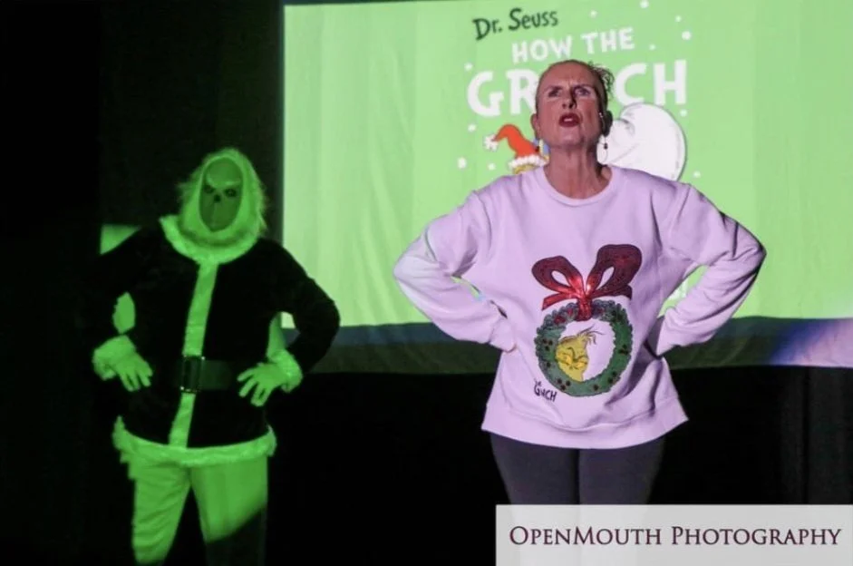 A woman and a person dressed as the Grinch stand in front of a green presentation or poster slide. The woman is wearing a white sweatshirt with a Christmas wreath and a bow design, and the Grinch is wearing a dark outfit with a green mask and gloves.