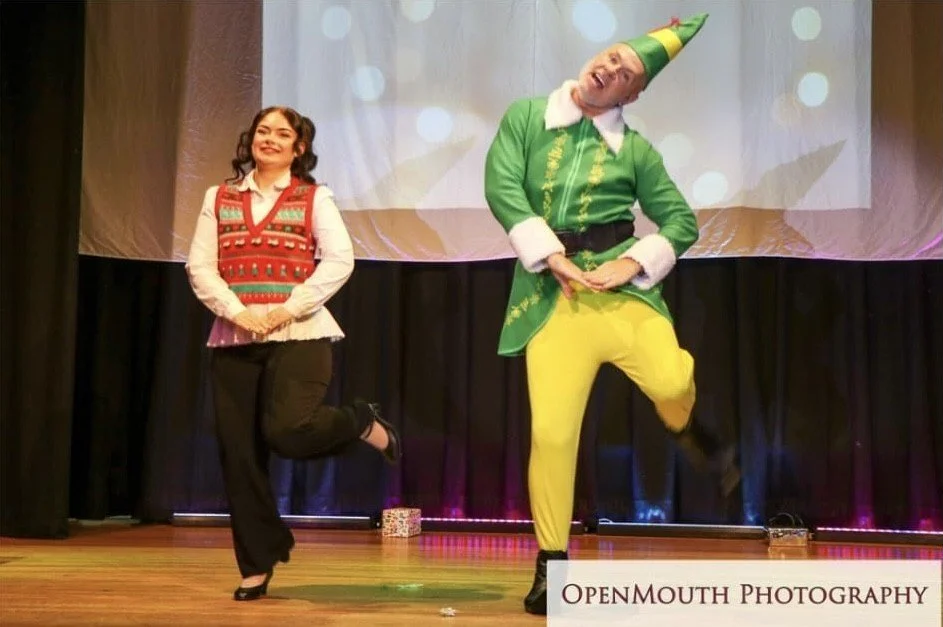 Two performers on stage dressed in holiday costumes, one as a woman in a festive sweater with long black hair, and another as an elf in bright green and yellow with a pointed hat, performing a dance.