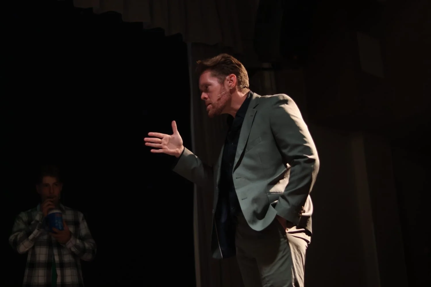 A man in a gray suit speaking passionately on stage, gesturing with his hand, while another person in the background drinks from a cup.