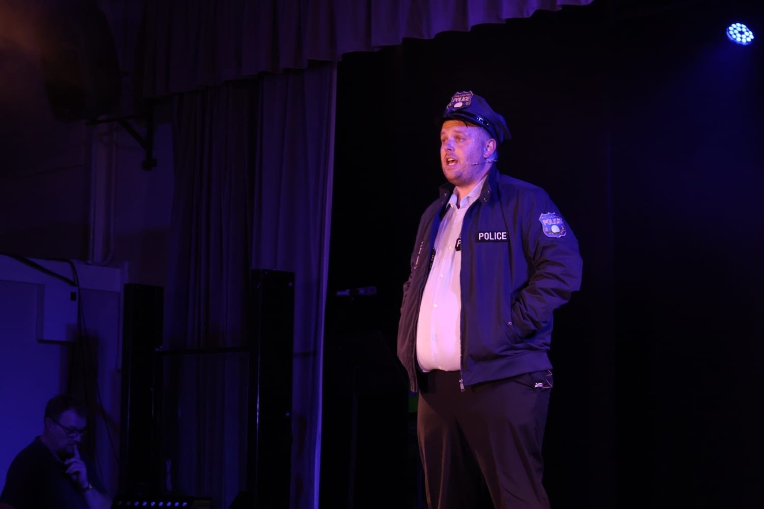 A man dressed as a police officer standing on stage with a microphone headset, speaking with his hands in his pockets. The background is dark with stage lighting.