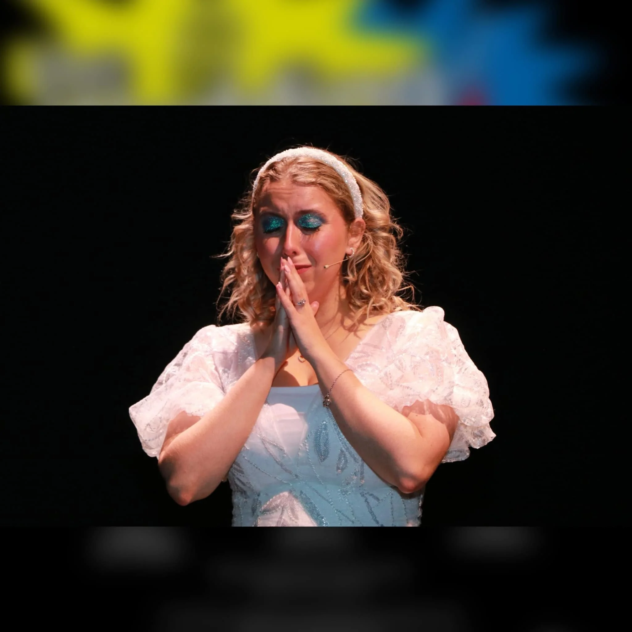 A woman with curly blonde hair in a white dress and glittery blue eye makeup, standing on stage with eyes closed and hands pressed together as if praying.