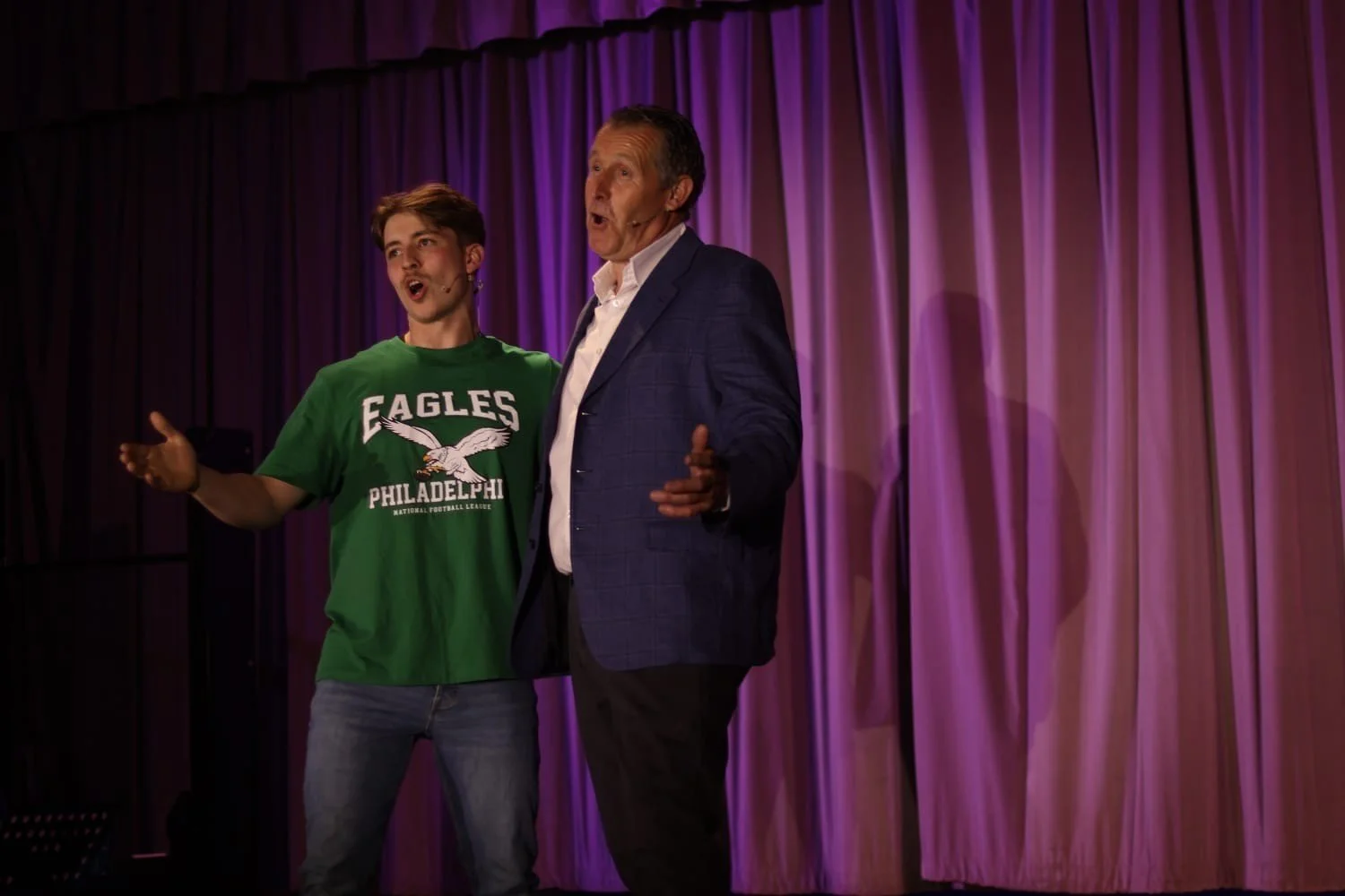 Two men singing on stage with purple curtains behind them. The younger man is wearing a green Eagles Philadelphia t-shirt, and the older man is wearing a blue checked blazer and white shirt.