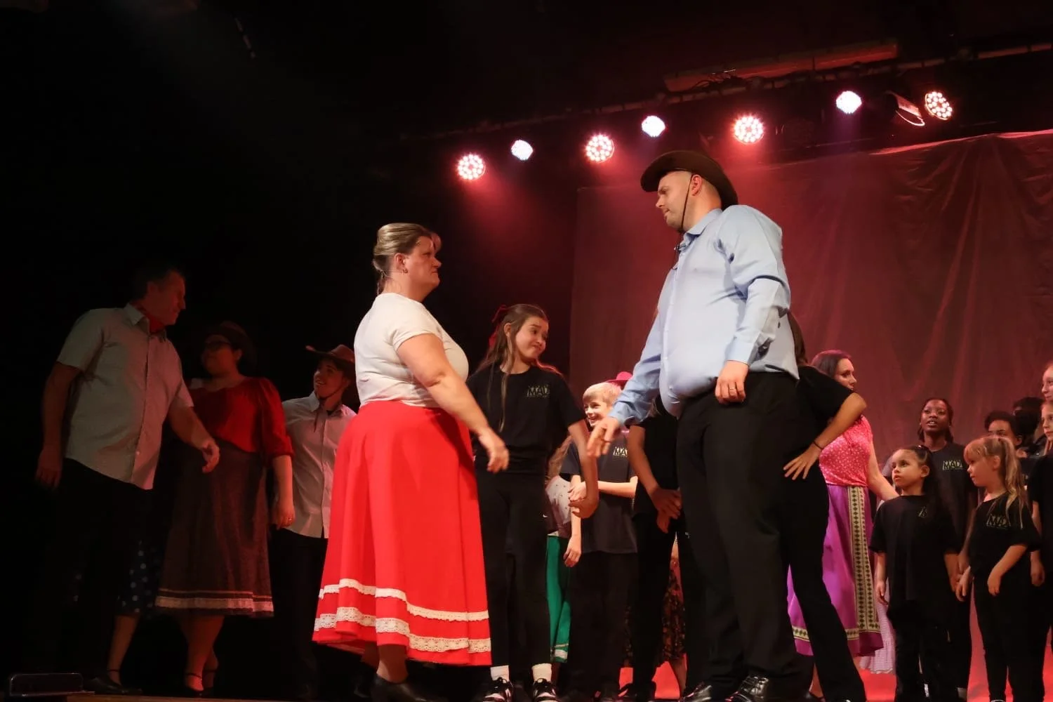 People on stage participating in a performance, with one woman in a red skirt and a man in a cowboy hat and blue shirt holding hands.