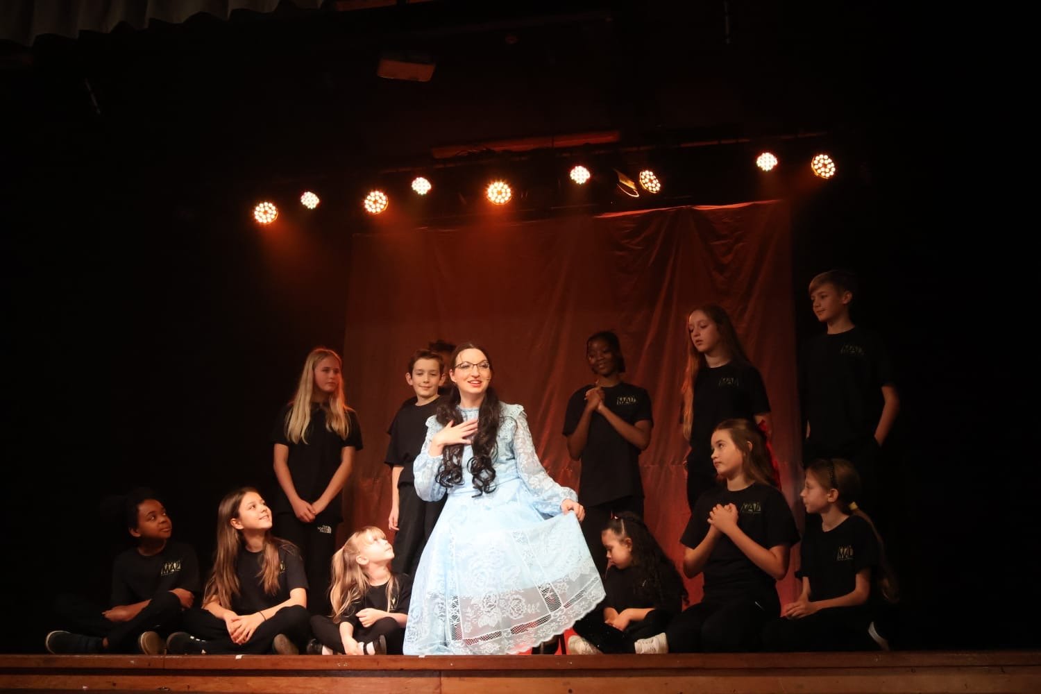 A woman in a light blue dress standing among children on stage during a performance or rehearsal, with children wearing black shirts around her, some sitting and some standing, against a dark background with stage lights overhead.