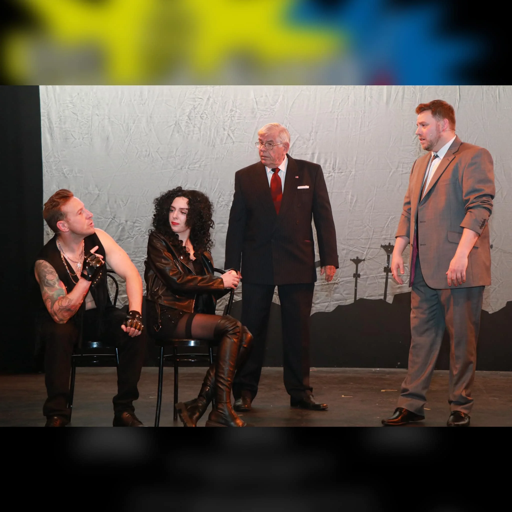 Four actors on stage during a play. Two are sitting, one is standing between them, and the other is standing to the right, wearing a suit. The background is a gray textured backdrop.