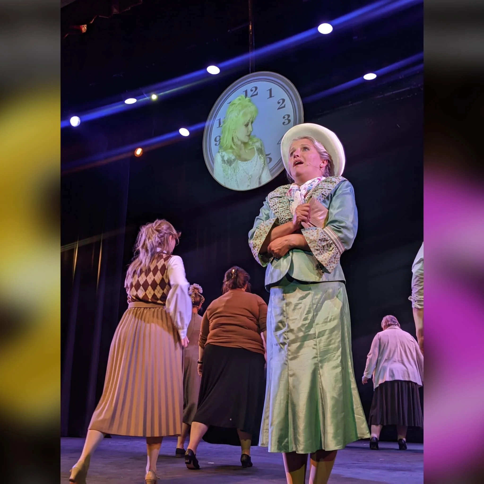 Stage scene with seven women, four facing away from the camera and three facing front, performing in a theatrical production. The woman in the front wears a wide-brimmed hat and a long green skirt, holding her hands near her chest. A large clock with