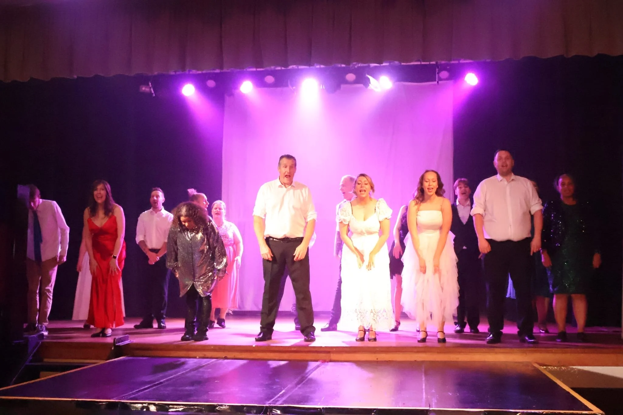 Group of performers on stage with purple lighting, dressed in formal and elegant attire, appearing to take a bow or conclude a performance.