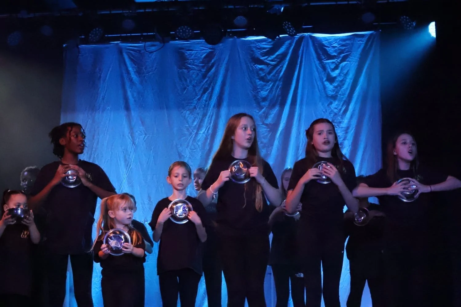 Group of young children performing on stage with a blue curtain backdrop, some holding silver bowls and singing.