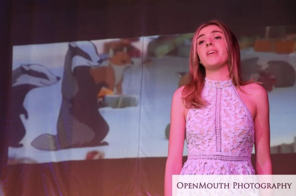 A young woman in a pink lace dress standing in front of a screen showing animated cartoon animals, including two penguins and a fox.