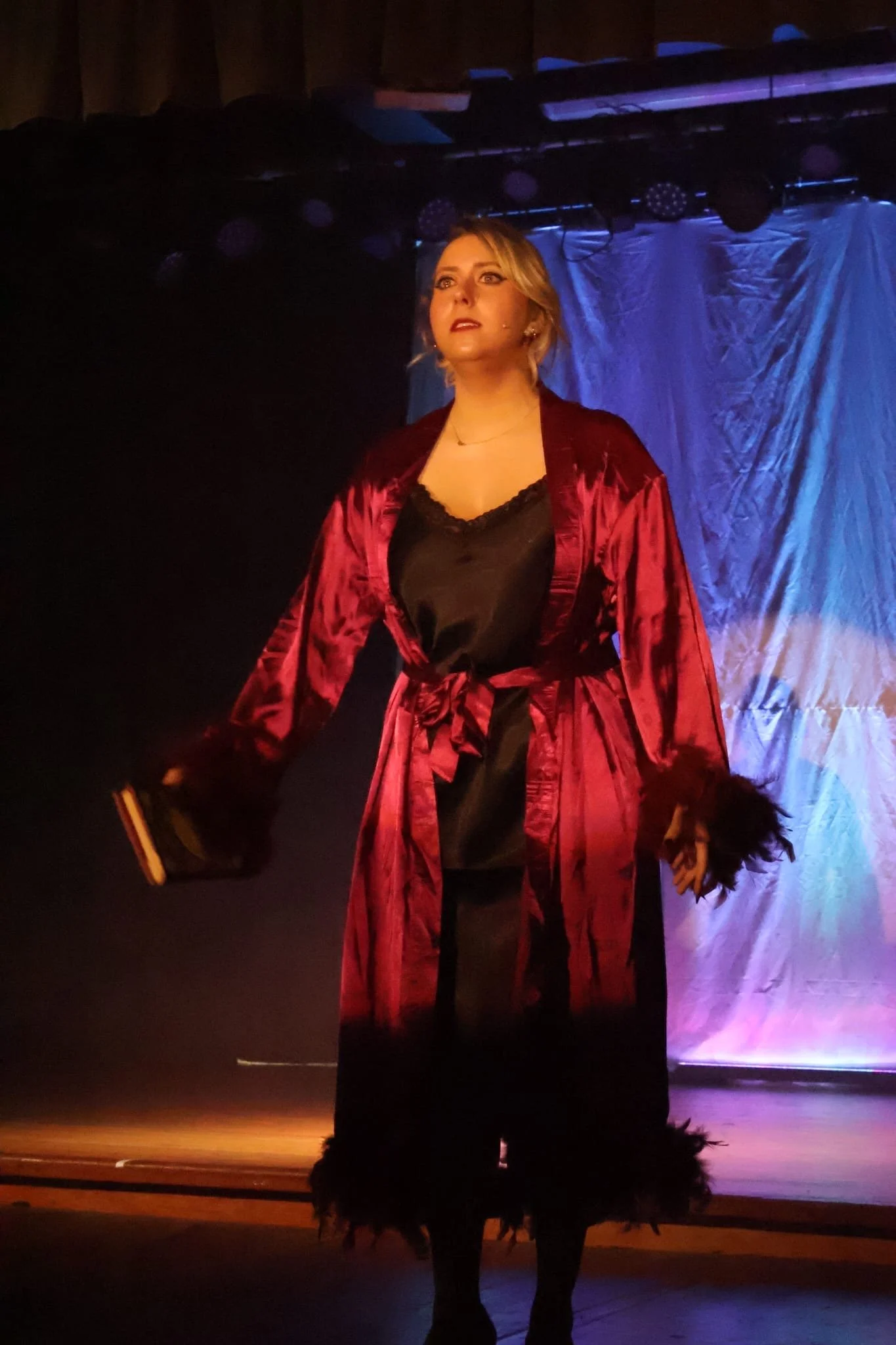 A woman standing on stage with dramatic lighting, wearing a black dress and a red satin robe with feathered cuffs and hem, holding a small object in her right hand.