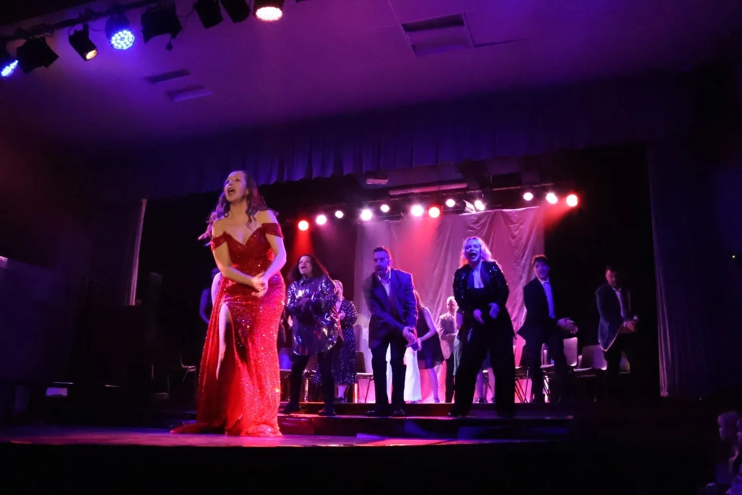 Performer in a red gown singing on stage with backup singers in black suits under colorful stage lights.