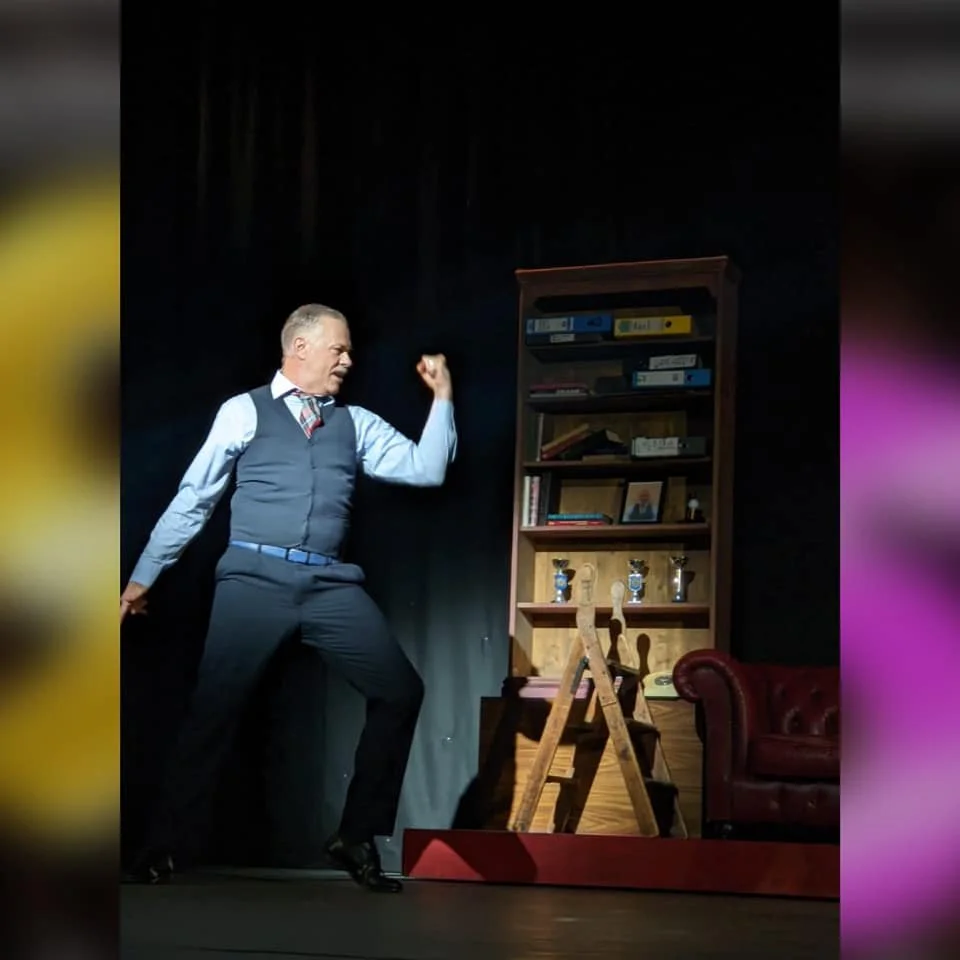 A man dressed in a vest, light blue shirt, and striped tie, striking a dynamic pose with his arm flexed, standing on a stage with a bookshelf, ladder, and a red armchair in the background.