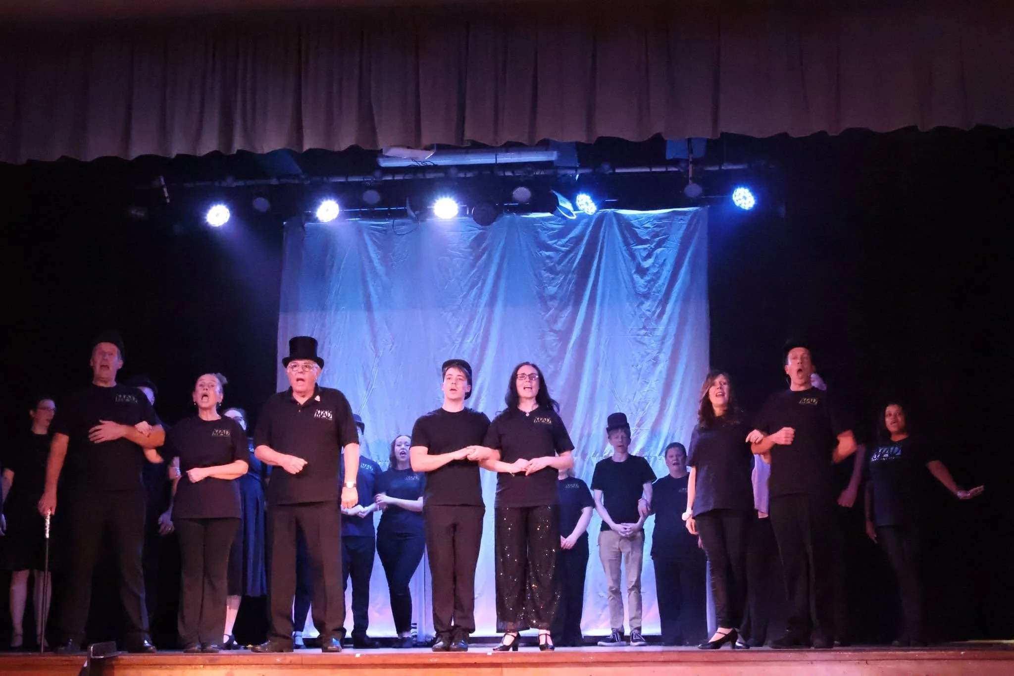A theatrical performance on stage with actors in black outfits, some in vintage attire, under blue stage lighting.