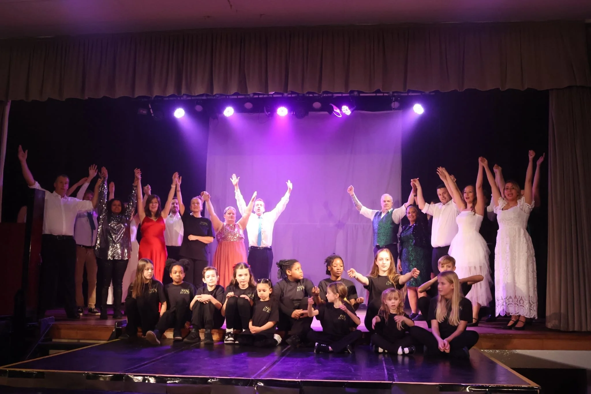 Group of children and adults on stage after a performance, some with arms raised, with a purple-lit backdrop and curtains.