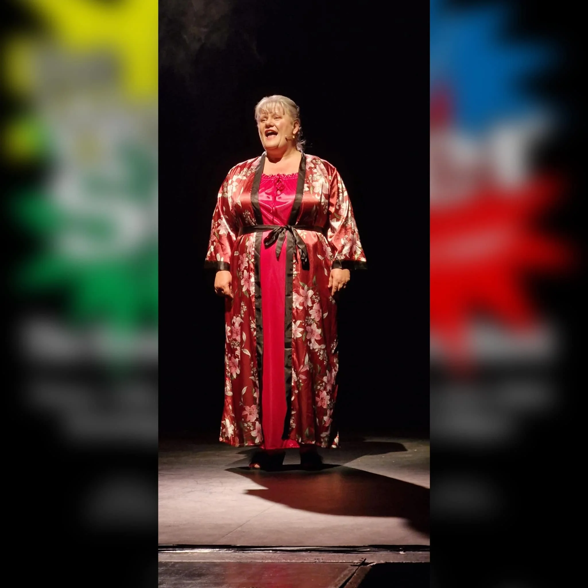 A woman singing on stage, wearing a pink and black floral kimono-style robe with a pink dress underneath.