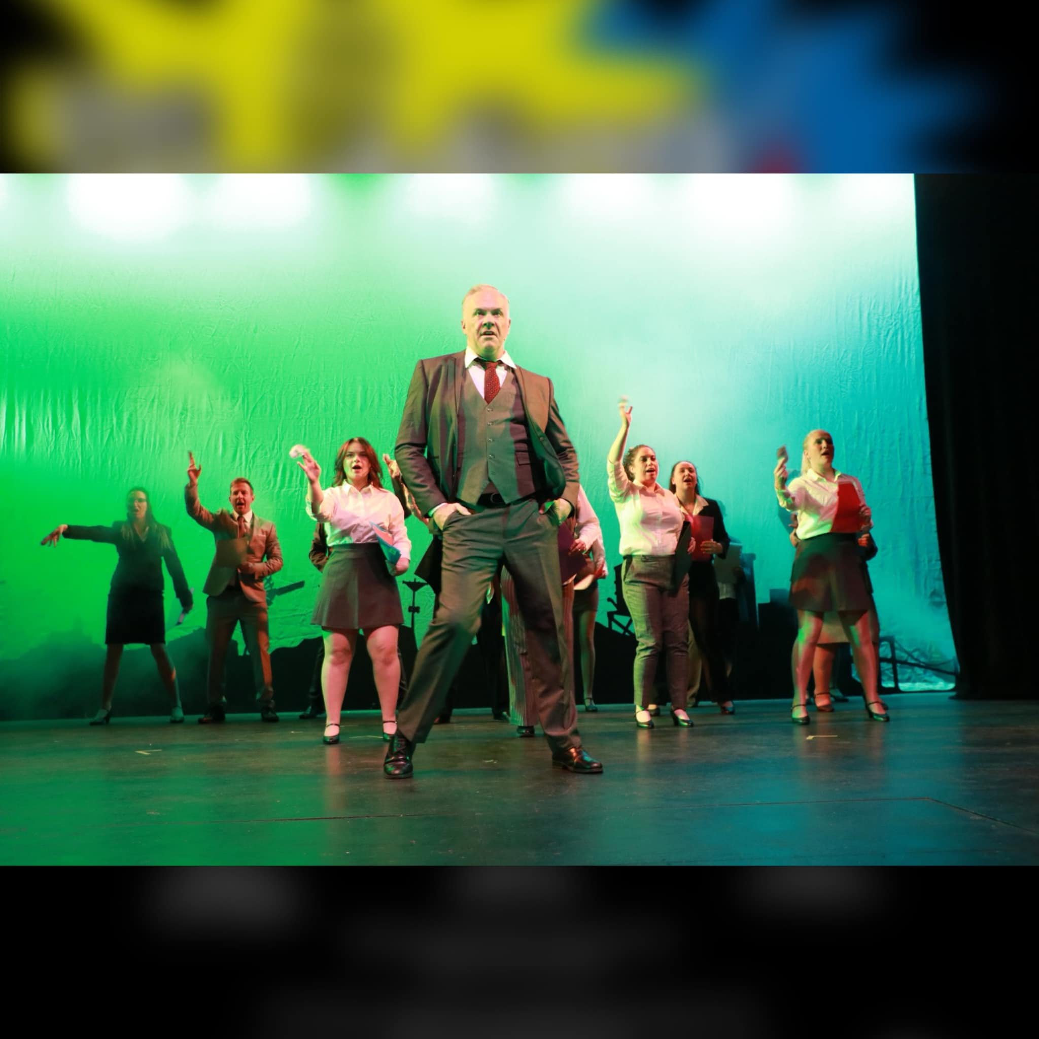 A theatrical performance with a man in a suit at the forefront surrounded by multiple women in business attire, on a stage with a large green and blue background.