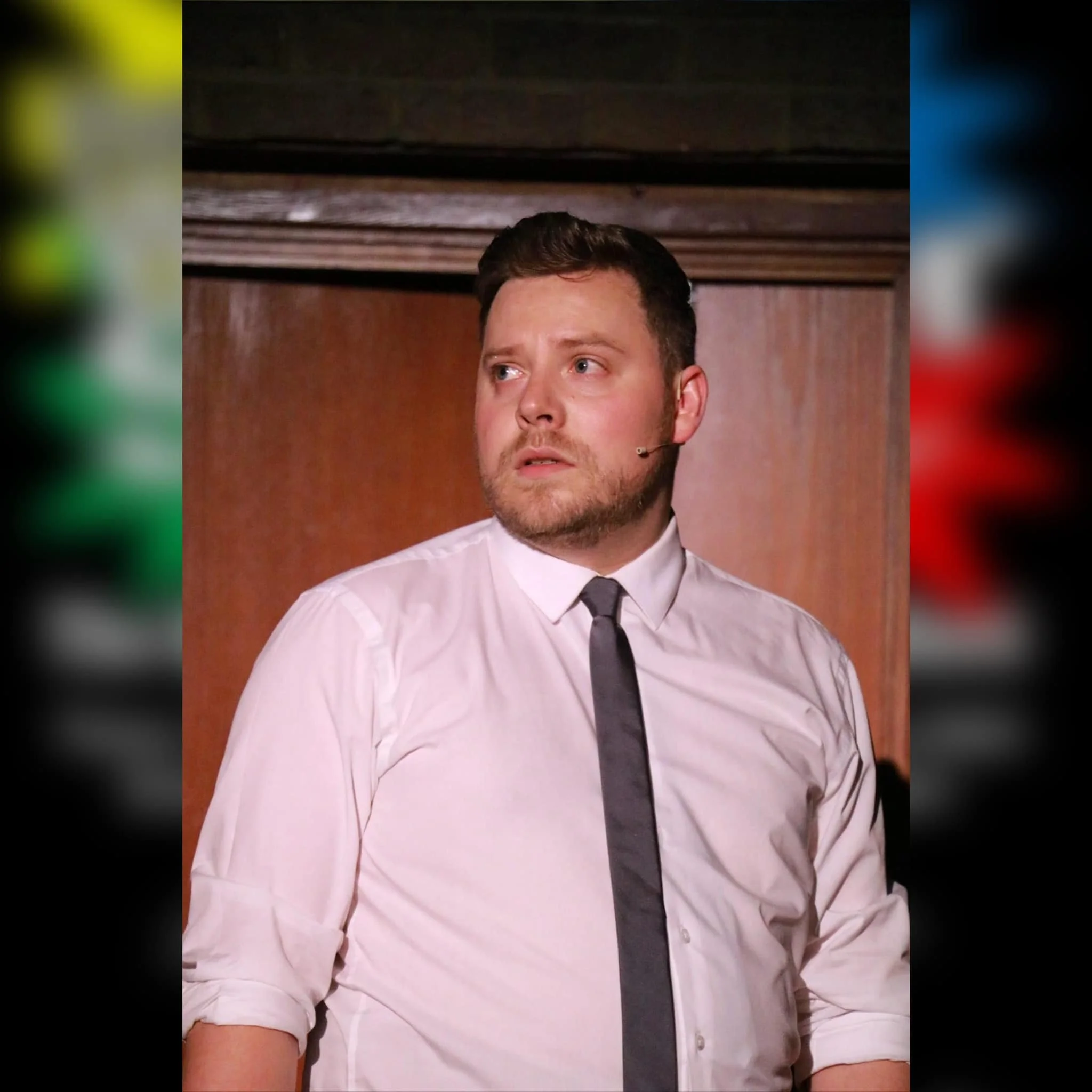 A man with short dark hair and a beard, wearing a white dress shirt and black tie, looks to the side with a surprised or concerned expression, standing in front of a wooden background.