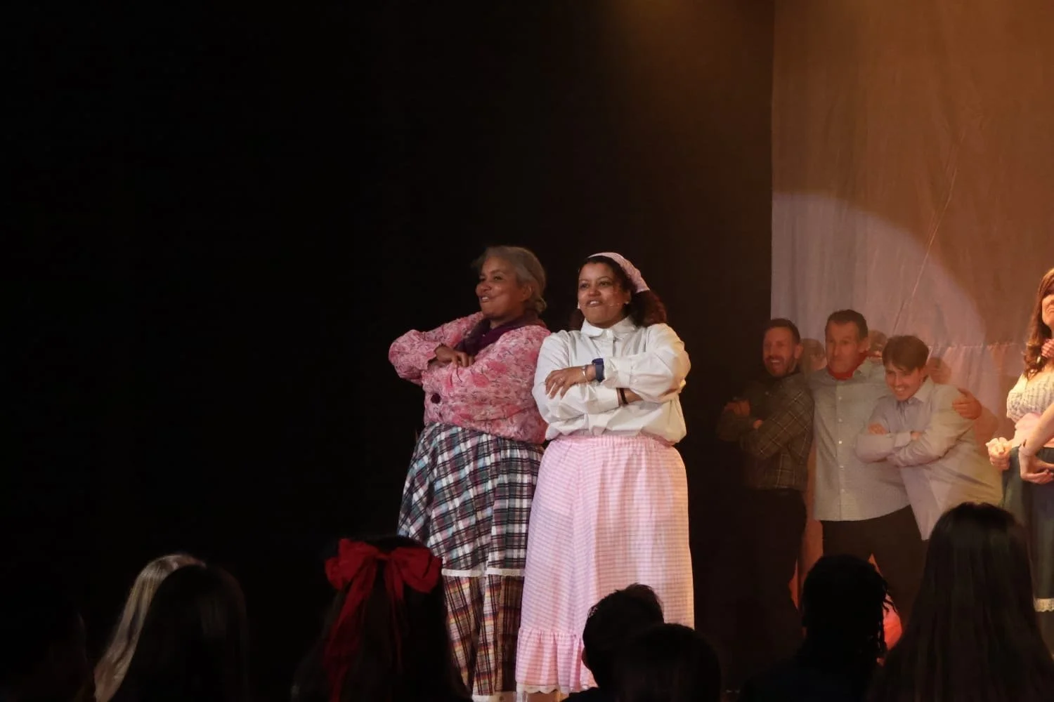 People standing on stage smiling with crossed arms dressed in casual and period costumes, during a performance or rehearsal in a theater.