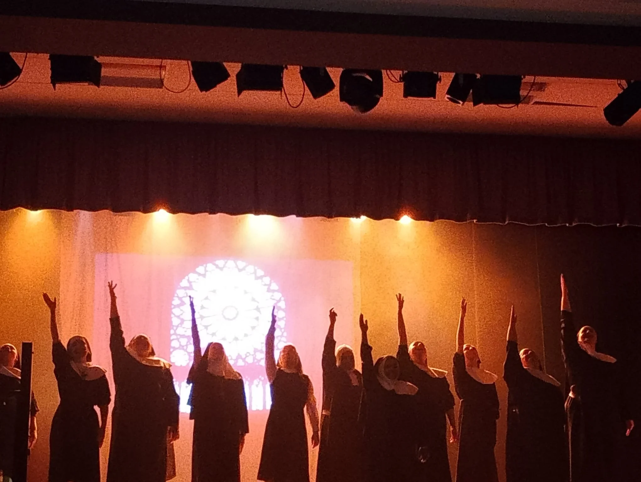A group of Nuns dressed in robes and habits on stage, raising one arm, with a stained glass window projected on the backdrop behind them.