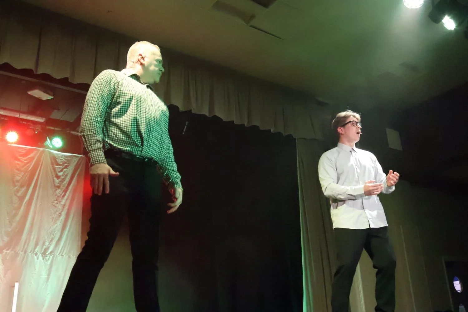 Two men performing on a stage under green and red lights; one wearing a checkered shirt and glasses, the other in a white shirt and glasses, with a dark background and curtains.
