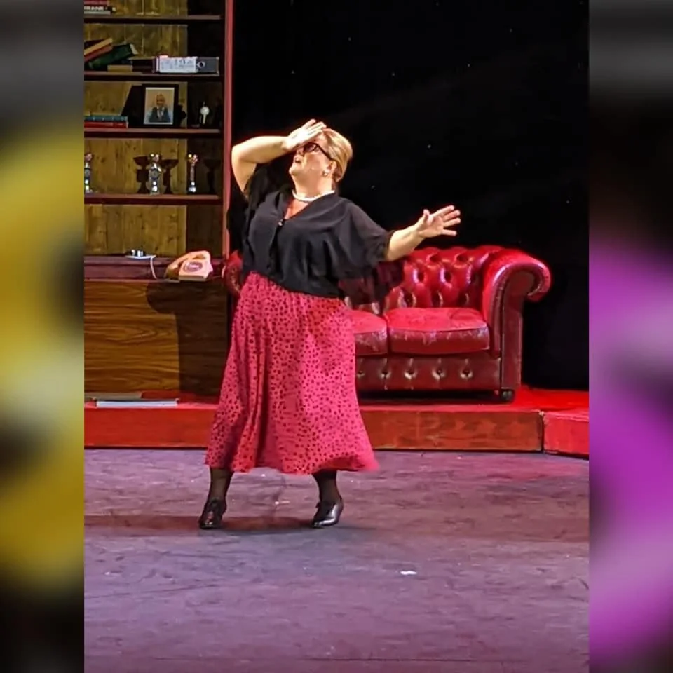 A woman on stage, dressed in a black top, red polka-dot skirt, and black heels, holding her hand to her forehead and extending her other arm outward, performing in front of a red Victorian-style sofa and a wooden bookshelf.