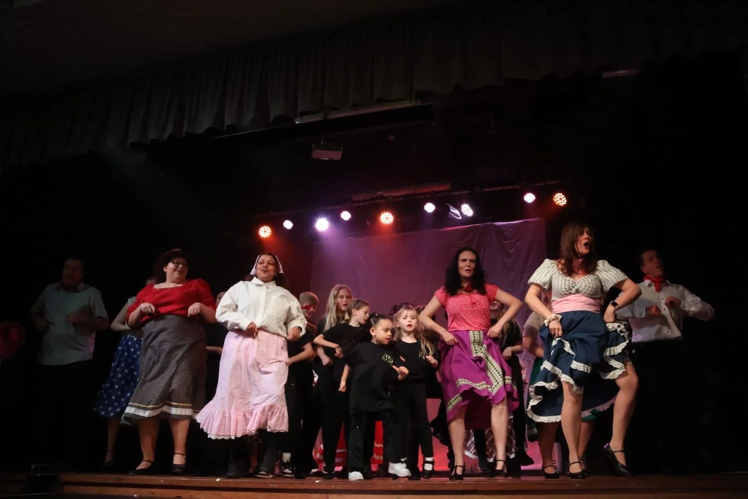 A group of children and adults on stage performing in a theatrical production, with colorful stage lights overhead.