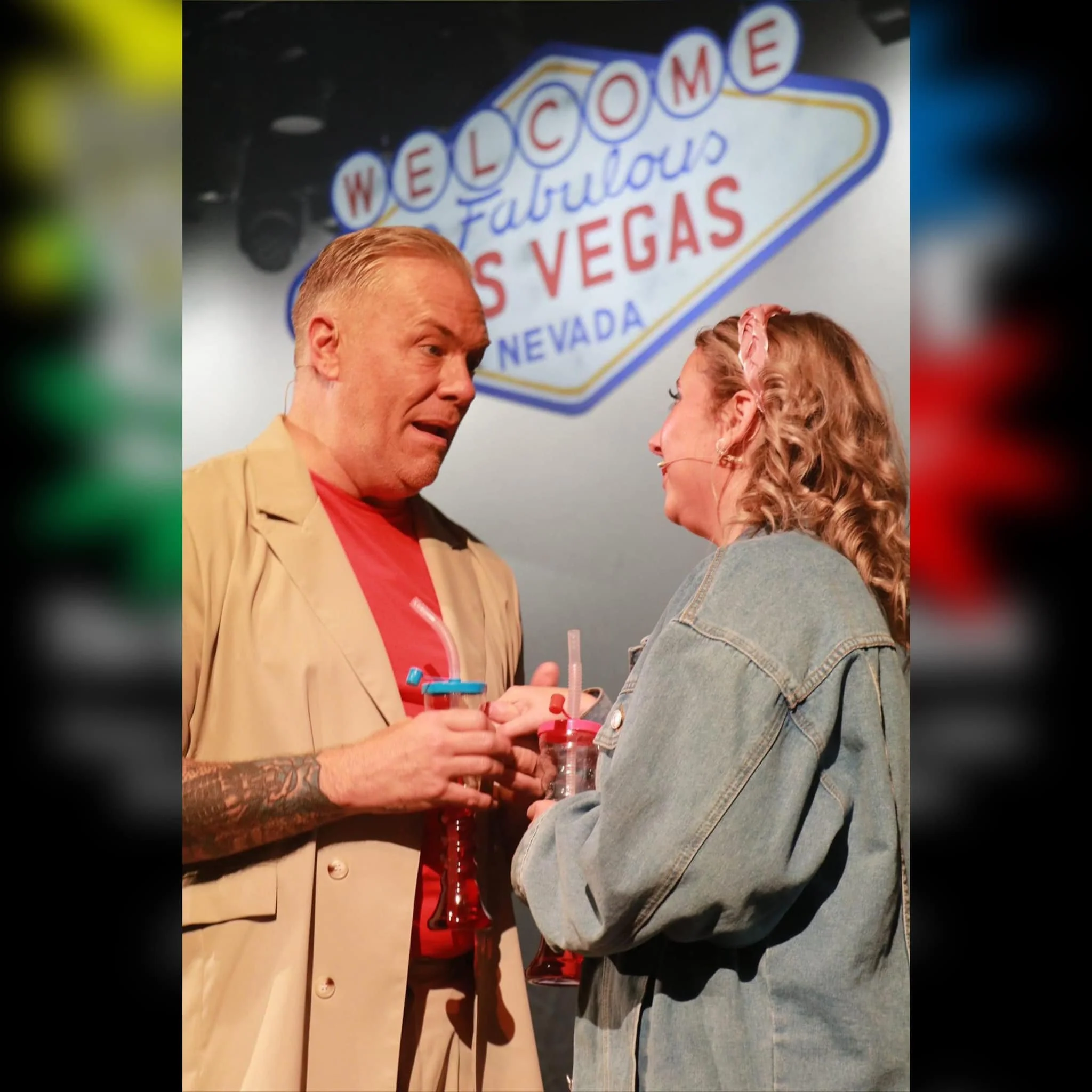 A scene with two people having a conversation in front of a 'Welcome to Fabulous Las Vegas Nevada' sign. One person, a man with short blond hair, wearing a beige jacket and red shirt, is holding a drink with a straw. The other, a woman with curly hai