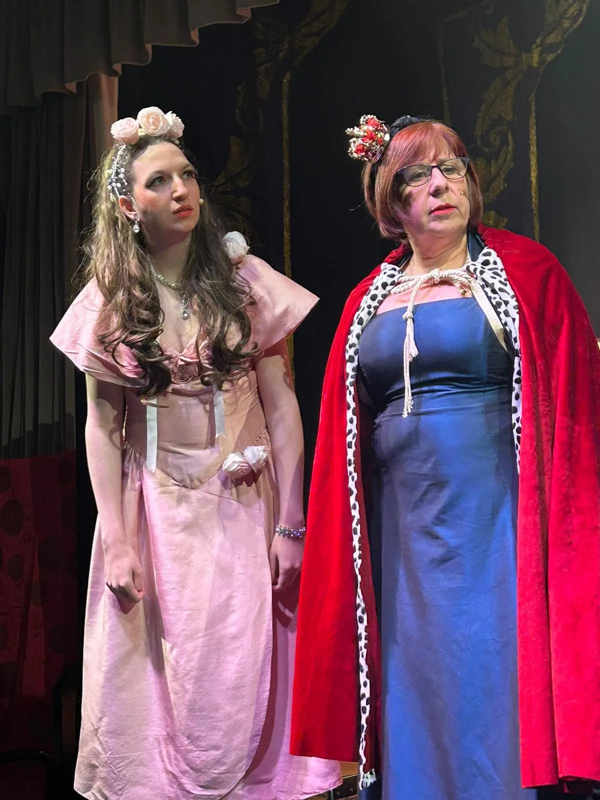 Two women dressed in theatrical costumes on stage, one in a pink dress with floral headpiece, the other in a blue dress with a red cape and black glasses, seen during a stage performance.