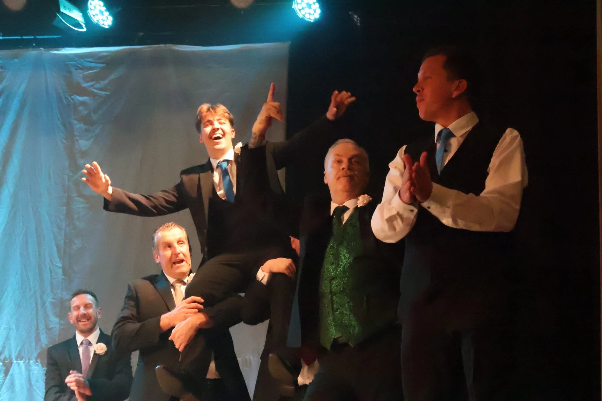 A group of five men dressed in suits and ties, performing on stage. One man is being lifted by another, all appearing joyful and animated, with a dark backdrop and stage lighting.