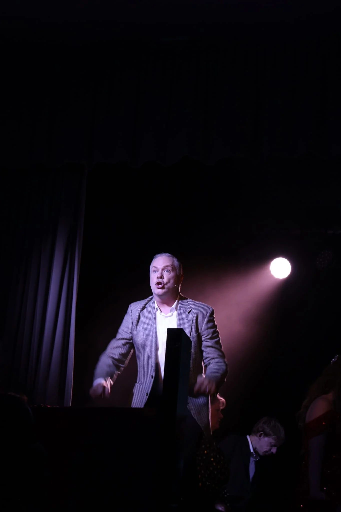 A man performing on stage, mid-sentence or singing, with spotlight behind him, in a dark theater setting.