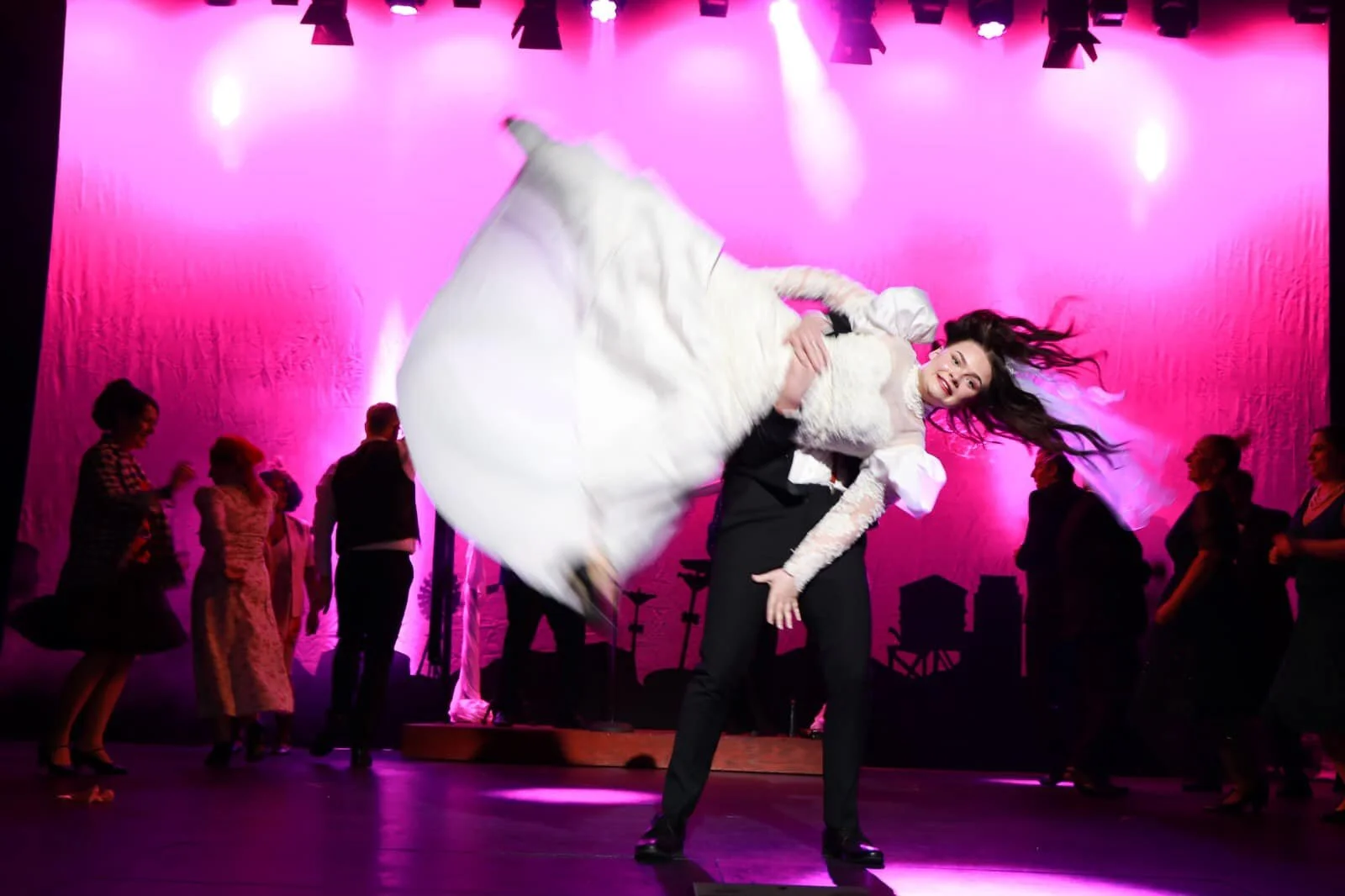 A woman in a vintage wedding dress being lifted into a cartwheel performing on stage with a group of people dancing around them, pink lighting in the background.