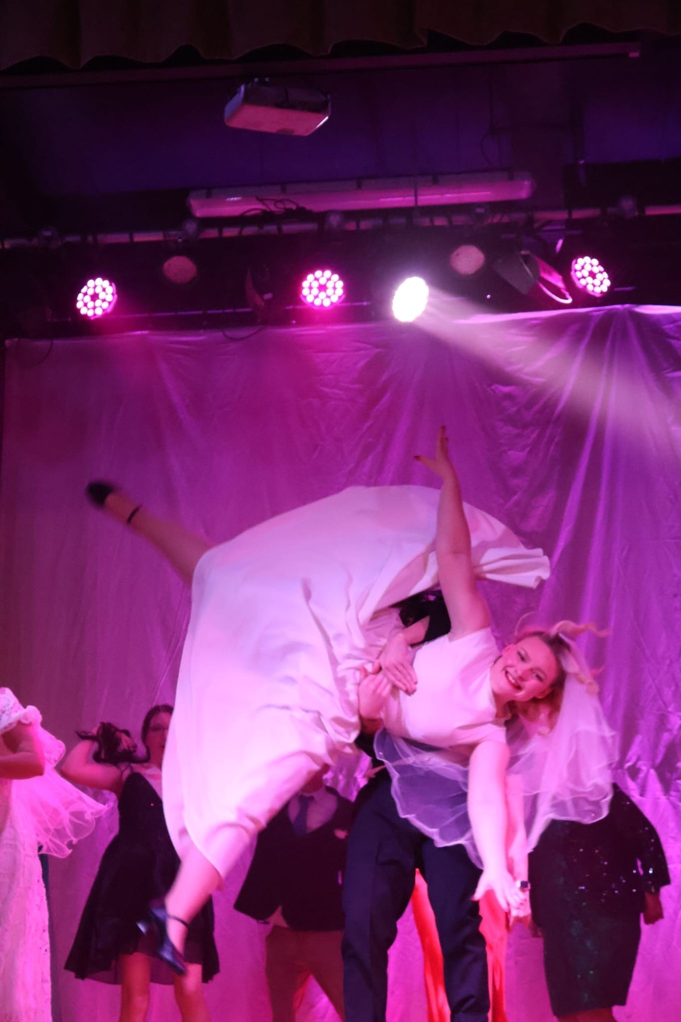 A woman in a white dress and veil performing an acrobatic pose on stage, surrounded by other performers, with pink stage lighting and a pink backdrop.