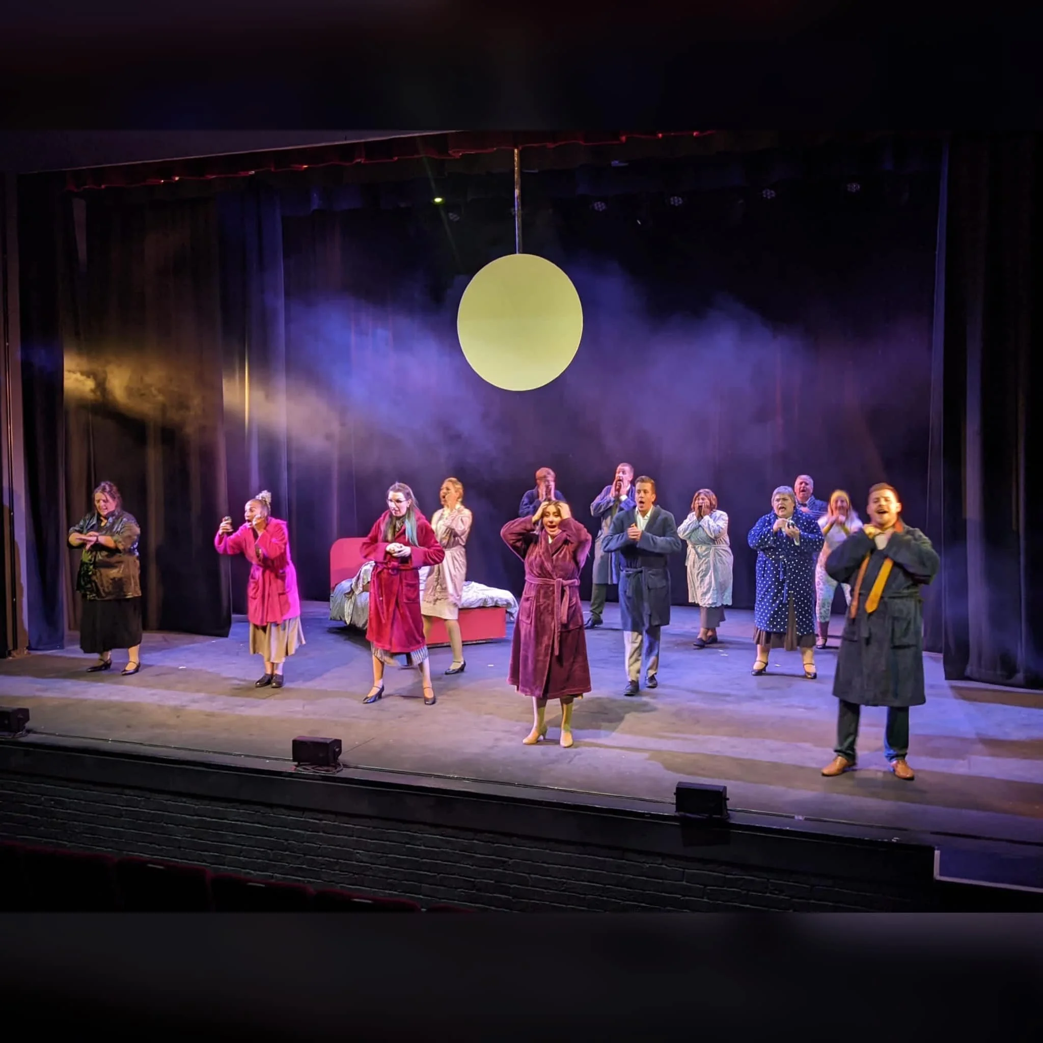 A theatrical stage with multiple actors in costumes performing a scene, with a pink bed and a large white circular object hanging above, and colorful lighting creating a dramatic atmosphere.