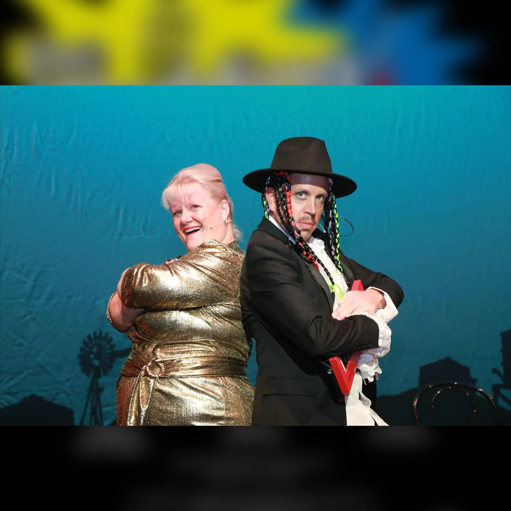 A woman in a glittery gold dress is smiling and standing with arms crossed, and a man in a black suit with a hat and colorful braids is standing with arms crossed.
