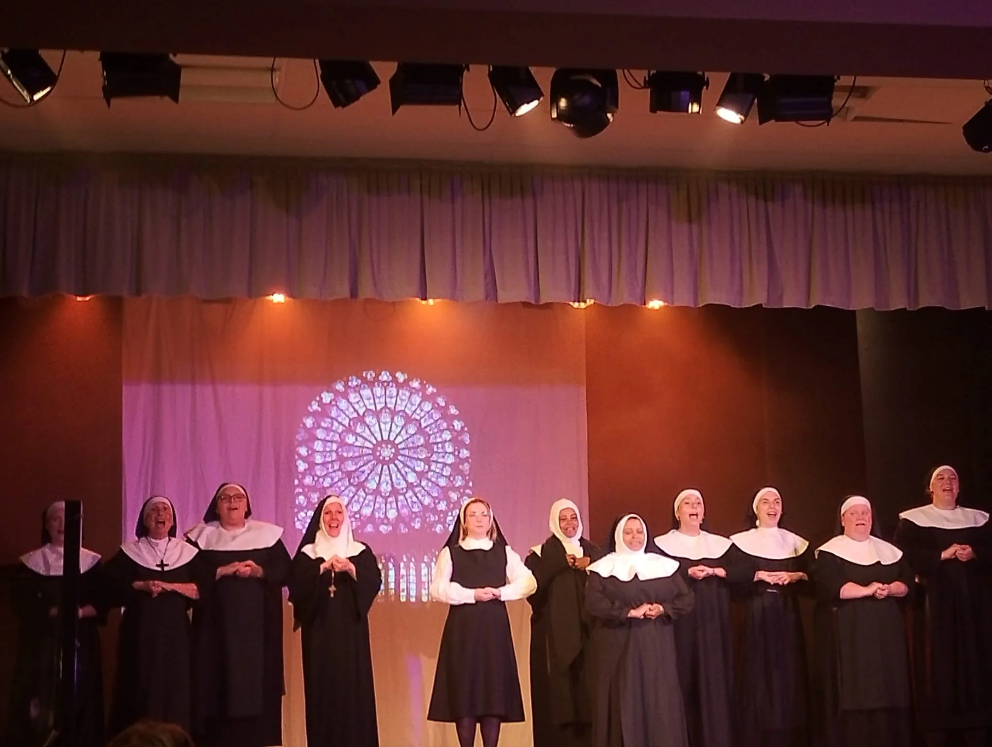 A group of women dressed as nuns singing on stage in front of a stained glass window projection.