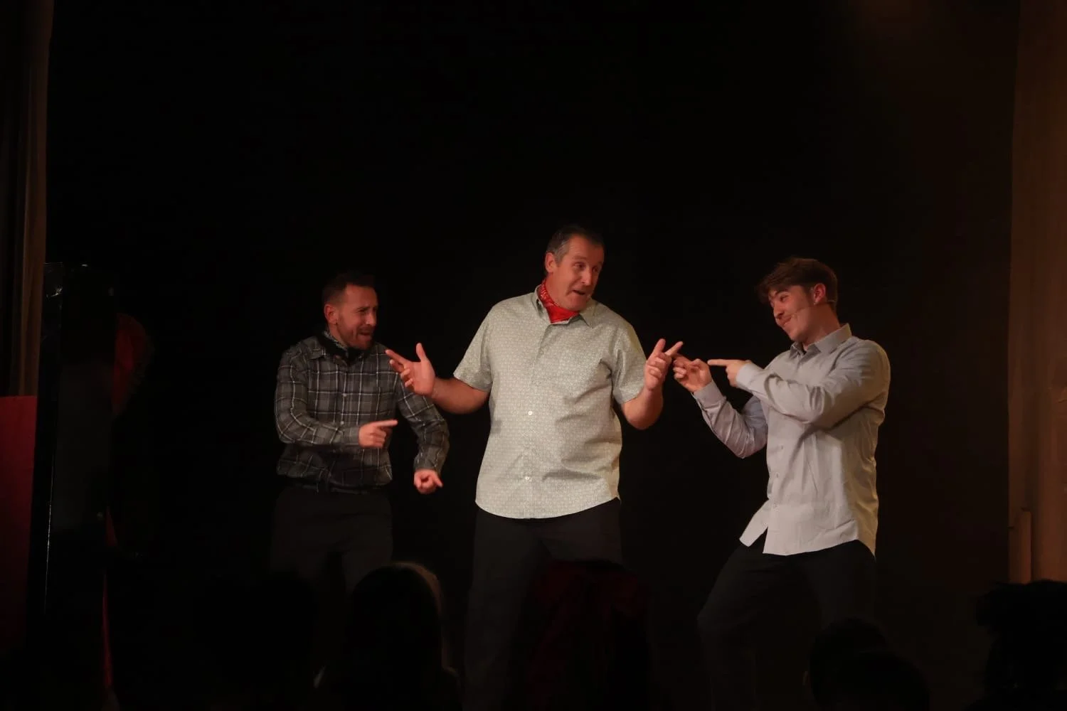 Three men performing on stage, two are smiling and one is gesturing with his hands, with a dark background.