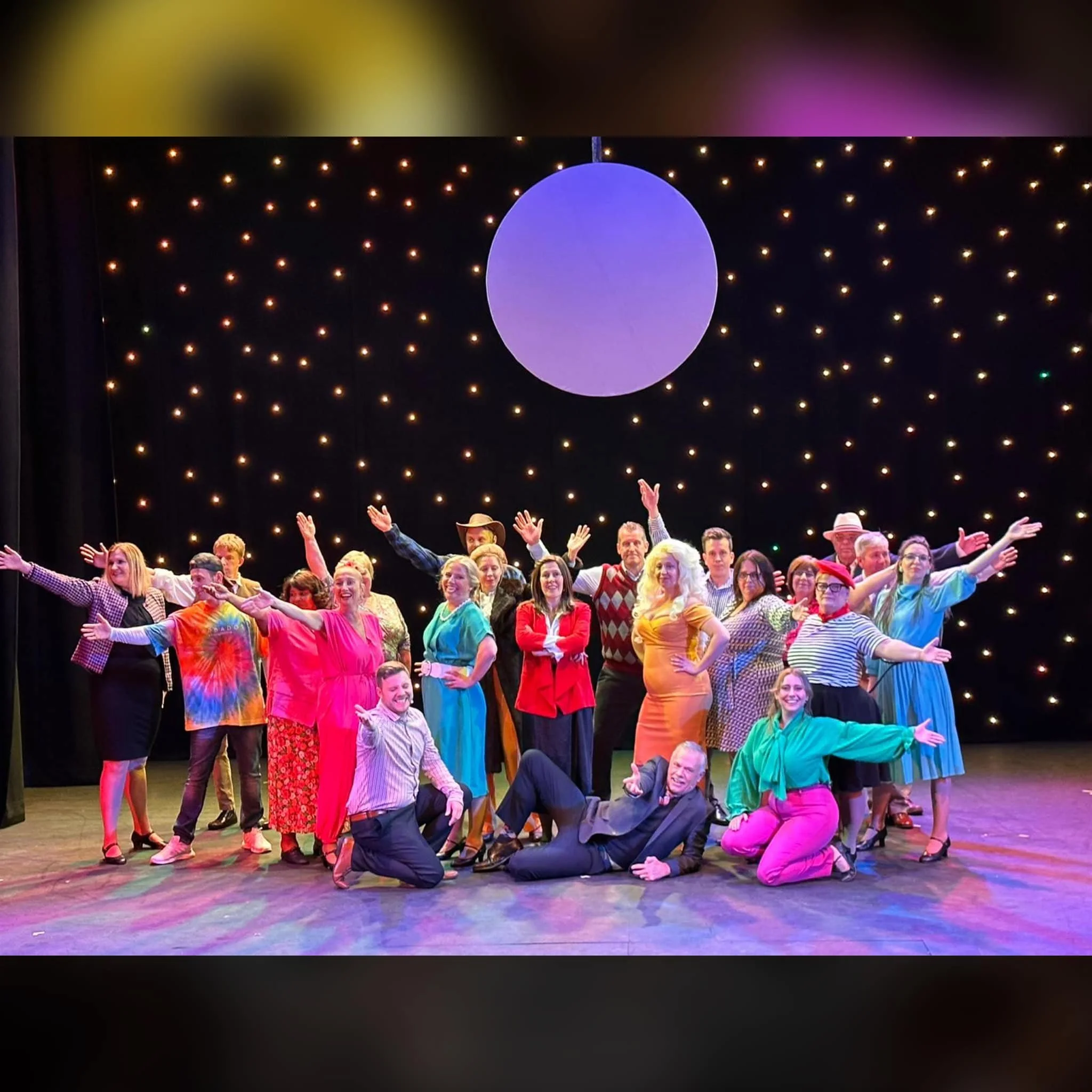 Group of performers on stage in colorful costumes, posing with arms outstretched and smiling, against a backdrop of black curtains with small twinkling lights and a large purple circular light.