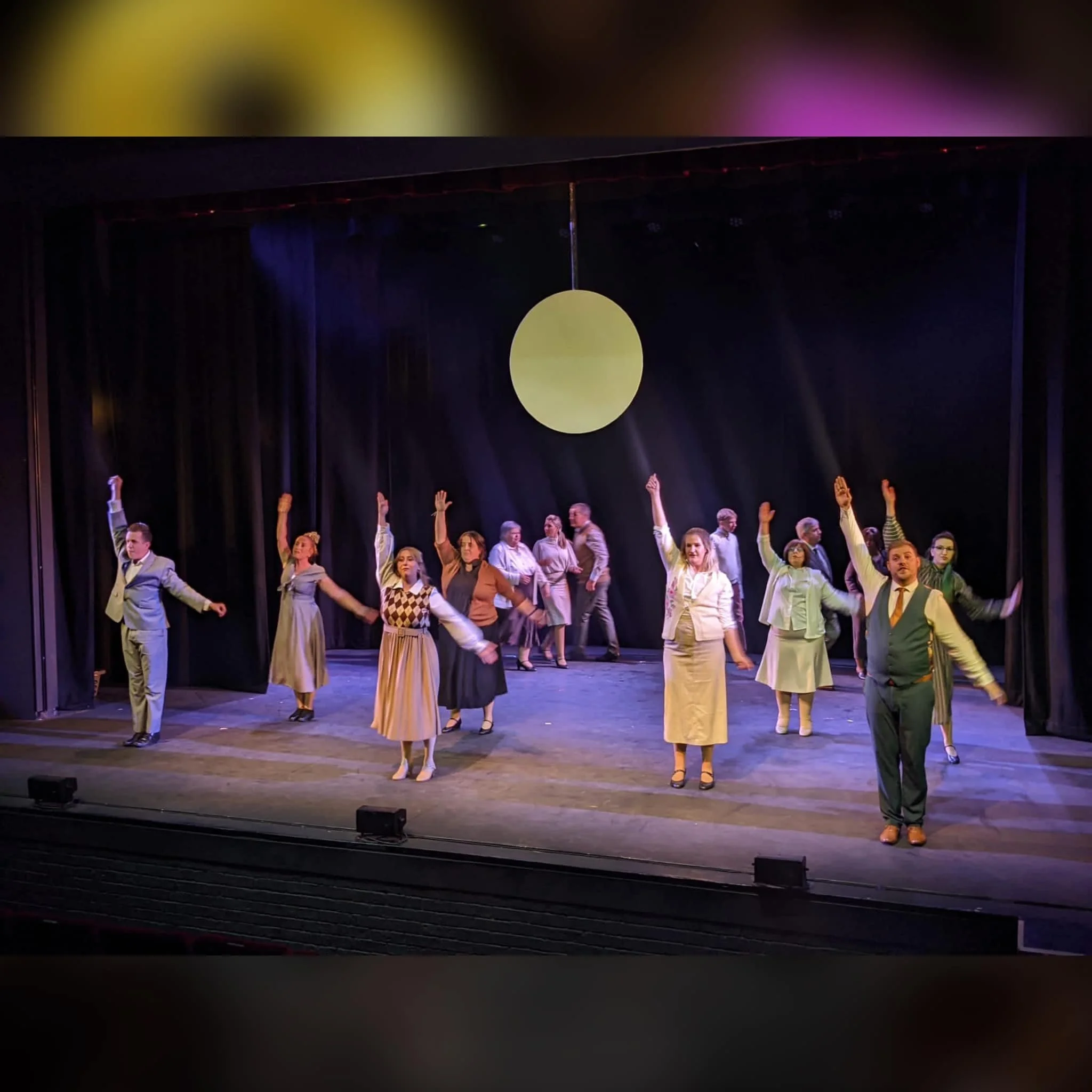 The image shows a group of performers on stage during a theater production, with some raising their hands, dressed in vintage clothing, in front of a black curtain and a large circular light or decoration hanging from the ceiling.