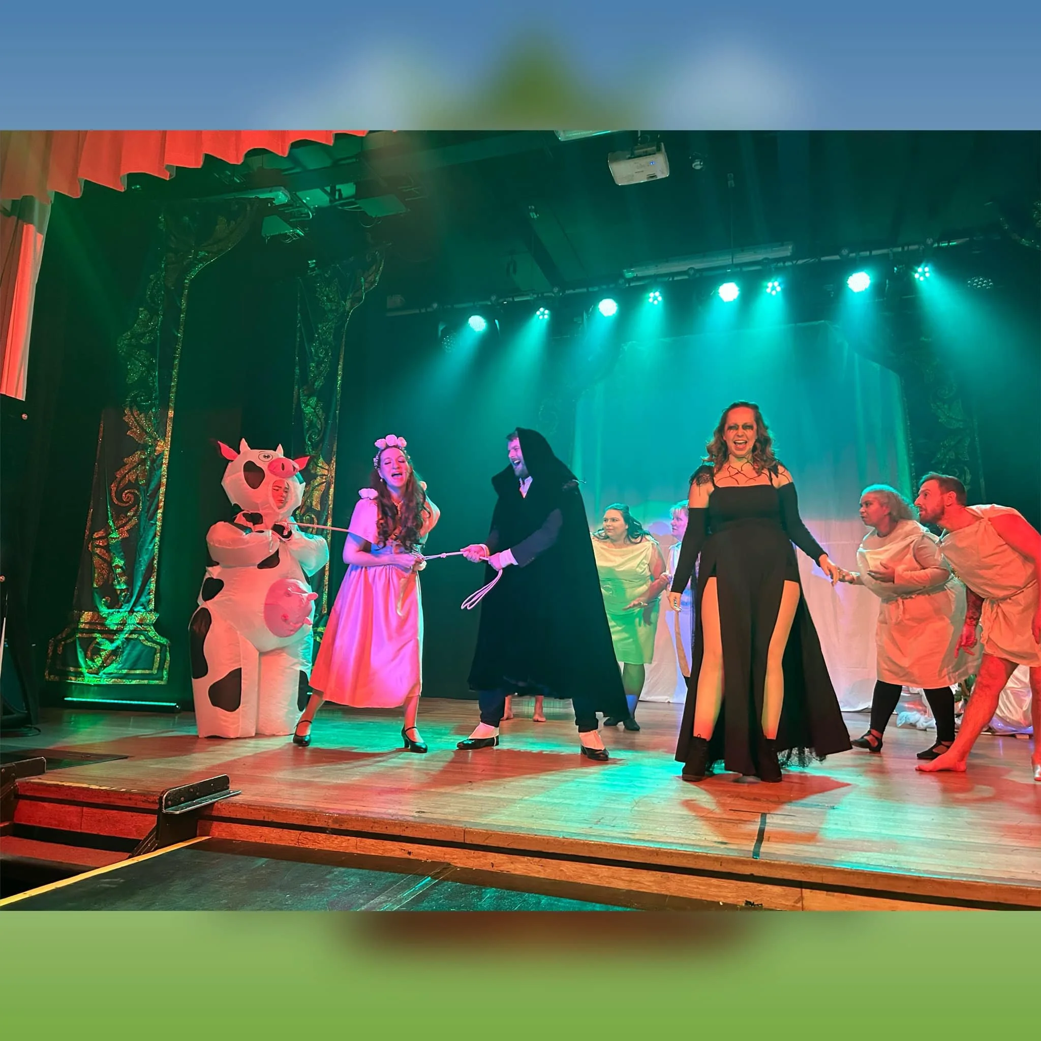 Actors perform on stage in a theatrical production. One actor wears a pig costume, while others are in colorful costumes, including a black dress, a pink dress, and costumes resembling medical personnel. The scene is lively with various characters in