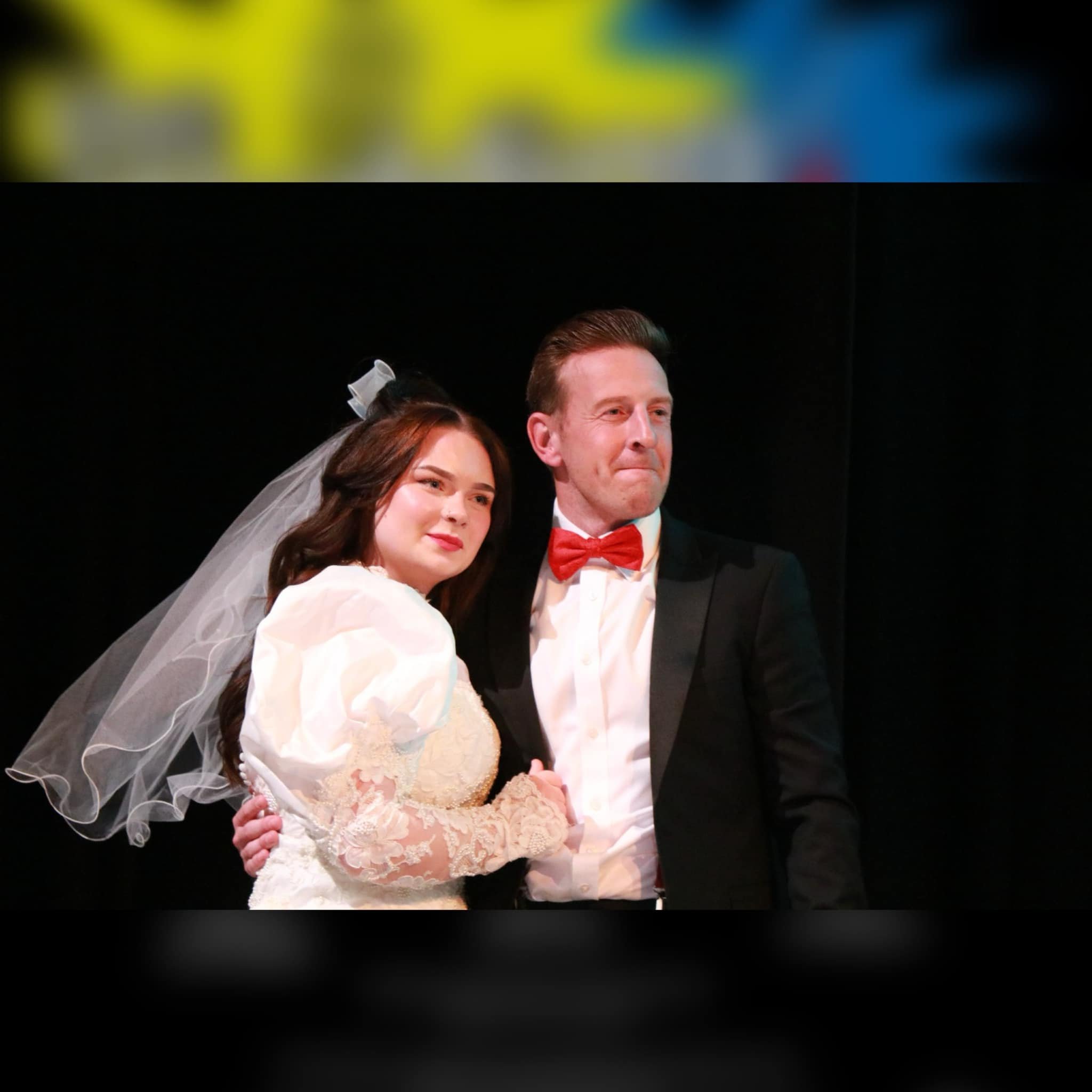 A man and woman in wedding attire, with the woman wearing a white dress and veil, and the man in a tuxedo with a red bow tie, standing close together in front of a dark background.