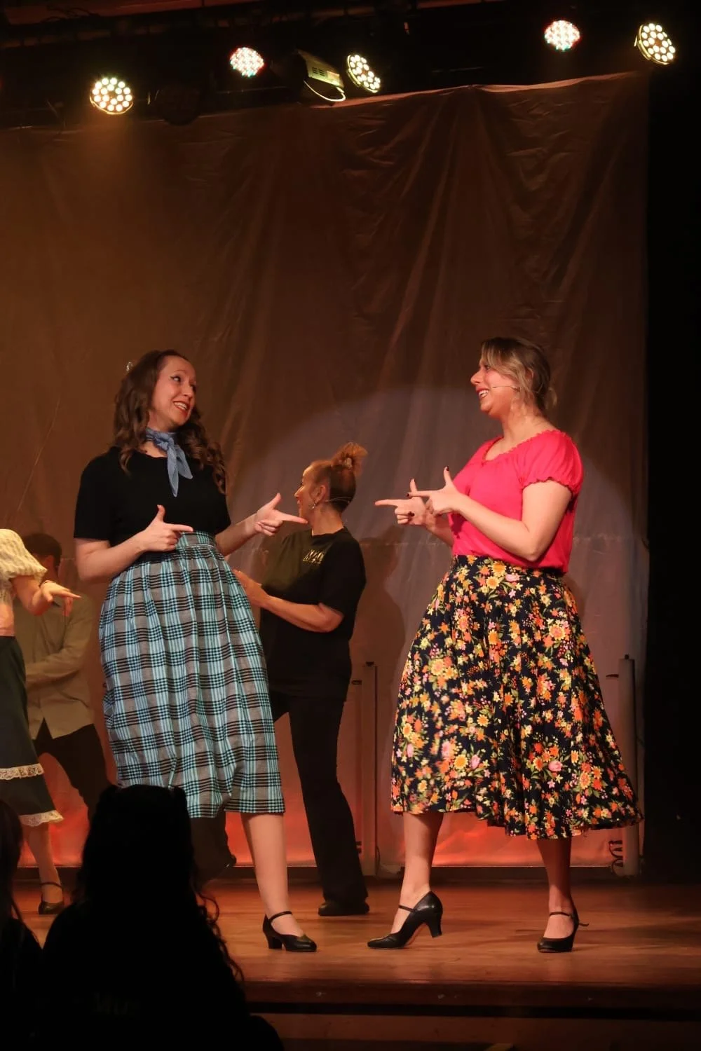 Two women on stage pointing at each other, smiling, during a theatrical performance with warm stage lighting and a curtain backdrop.