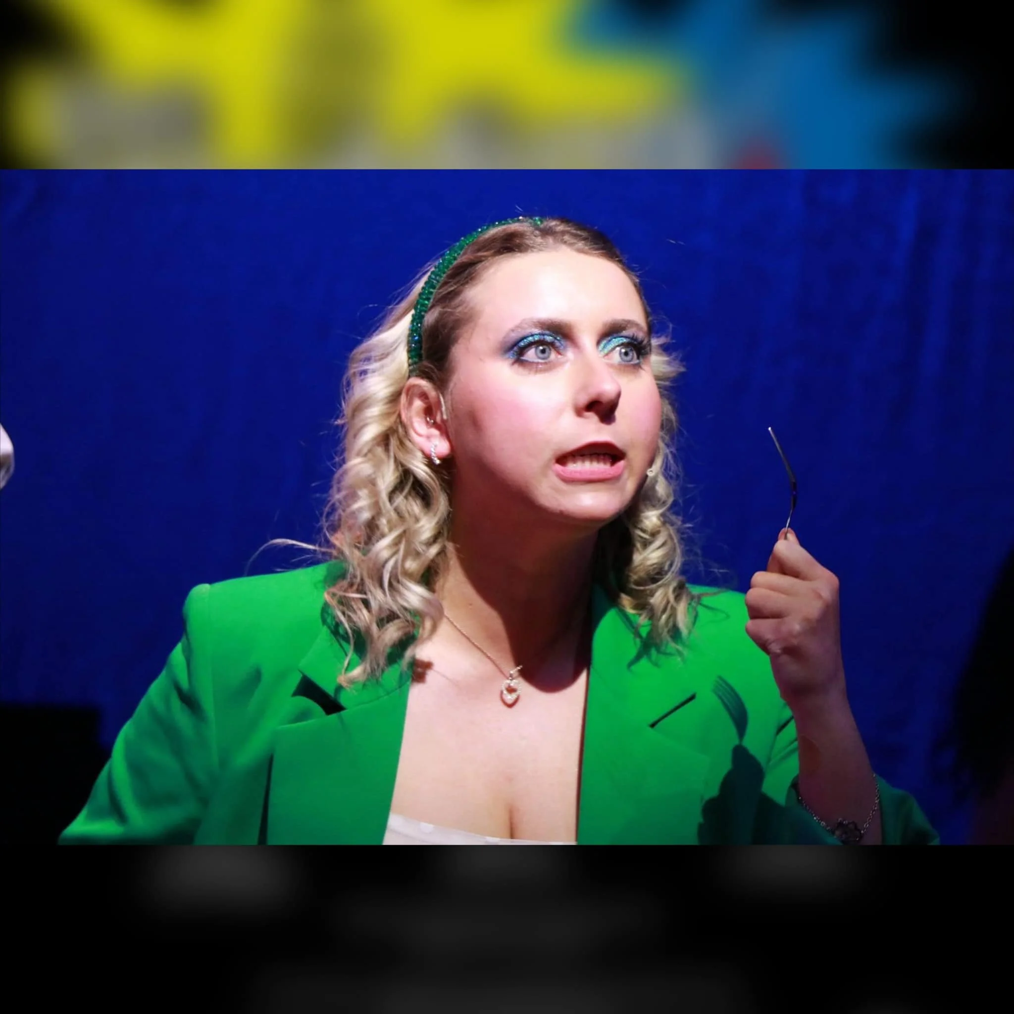 A woman with blonde curly hair, wearing a green blazer and a hairband, holding eyeglasses in her right hand, with a surprised or confused expression, standing against a blue background.