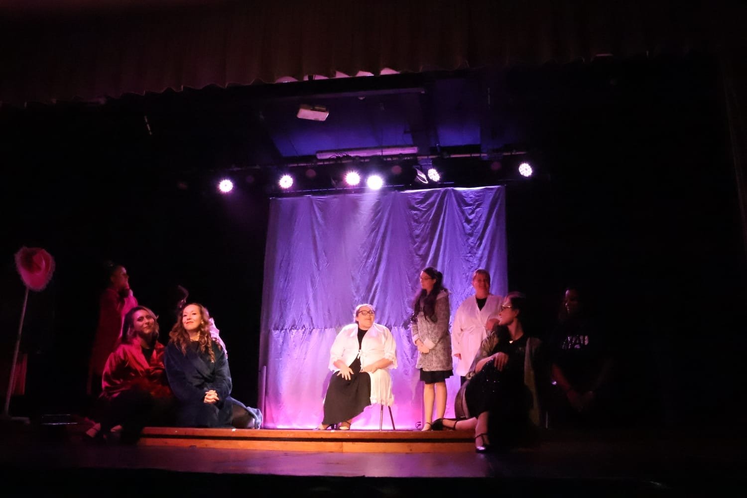 Theater cast performing on stage with purple lighting and a backdrop, five actors sitting and standing in costumes during a play.