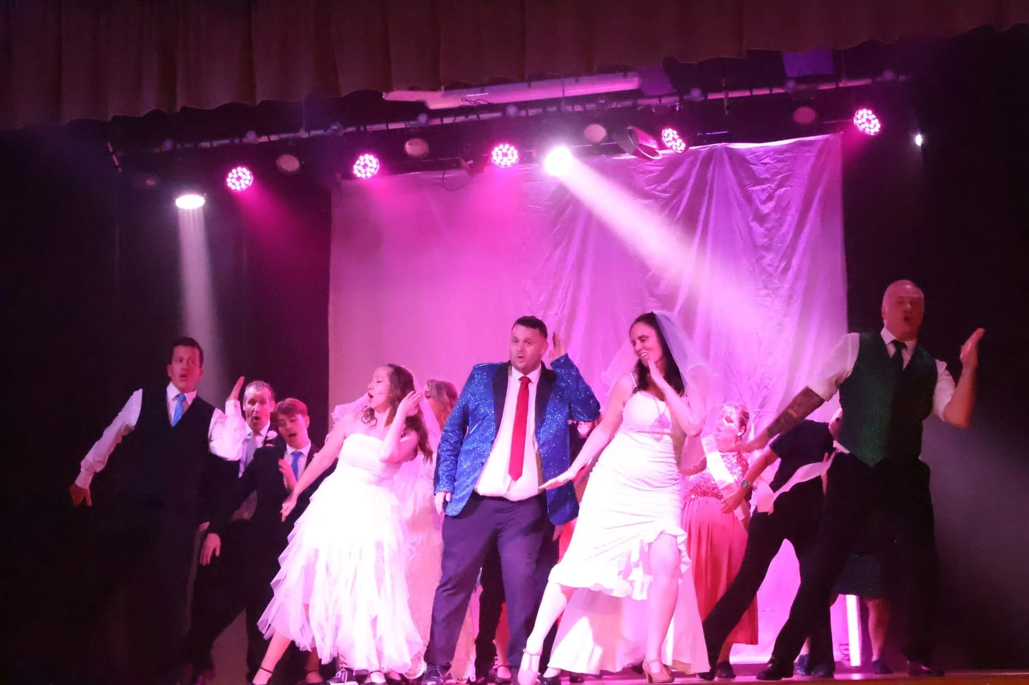 Stage performance with several actors, some in wedding and formal attire, under purple and pink stage lighting, with a pink backdrop.