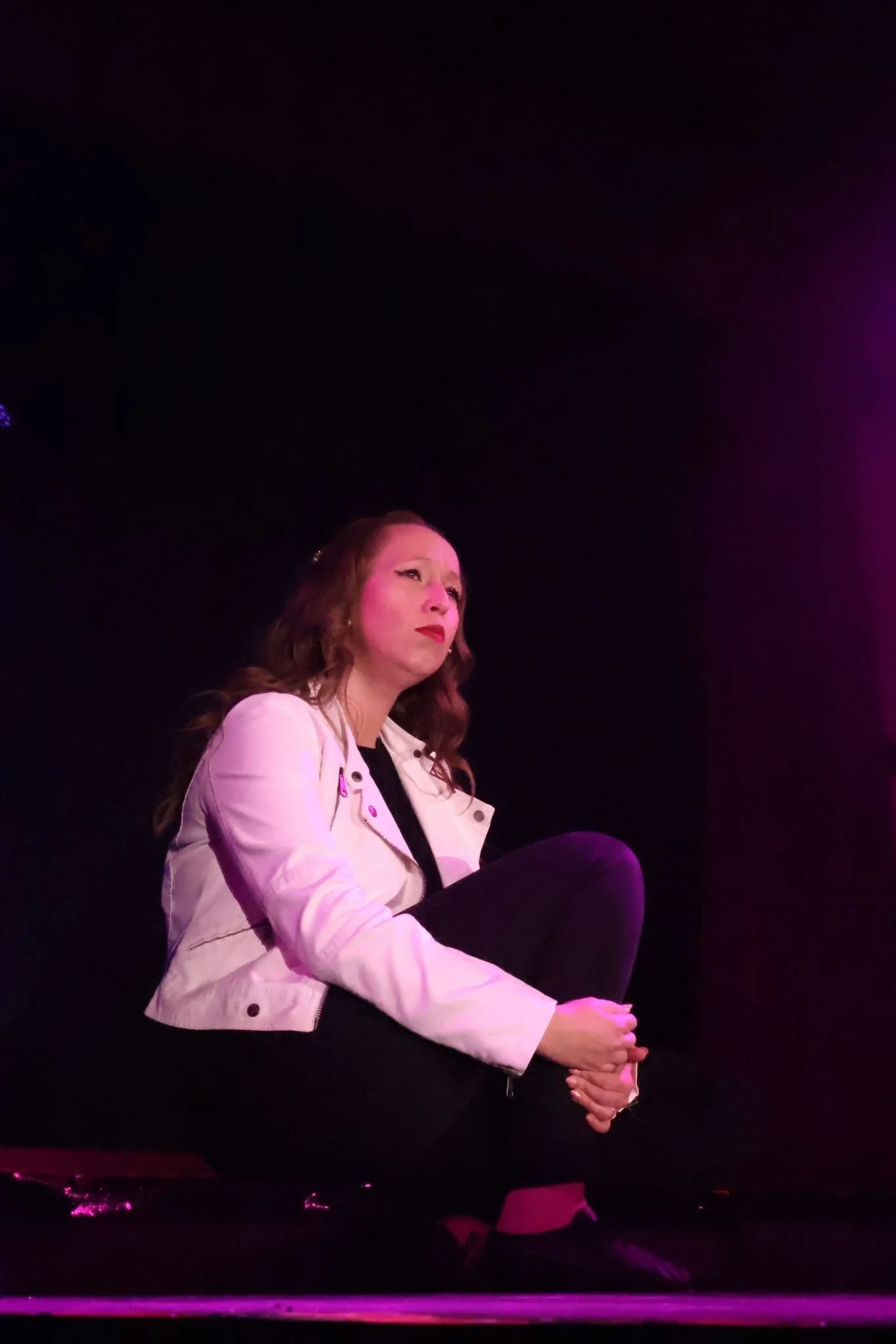 A woman with wavy brown hair sitting on the floor with black pants, a white jacket, and black shoes, looking thoughtful against a dark background with pink lighting.
