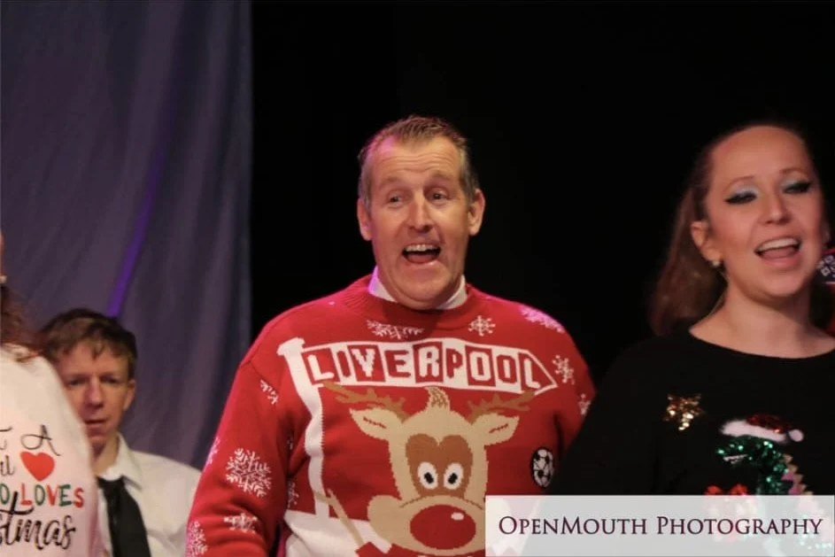 People singing on stage, with one man wearing a Christmas sweater with a reindeer and 'LIVERPOOL' written on it, and a woman next to him.