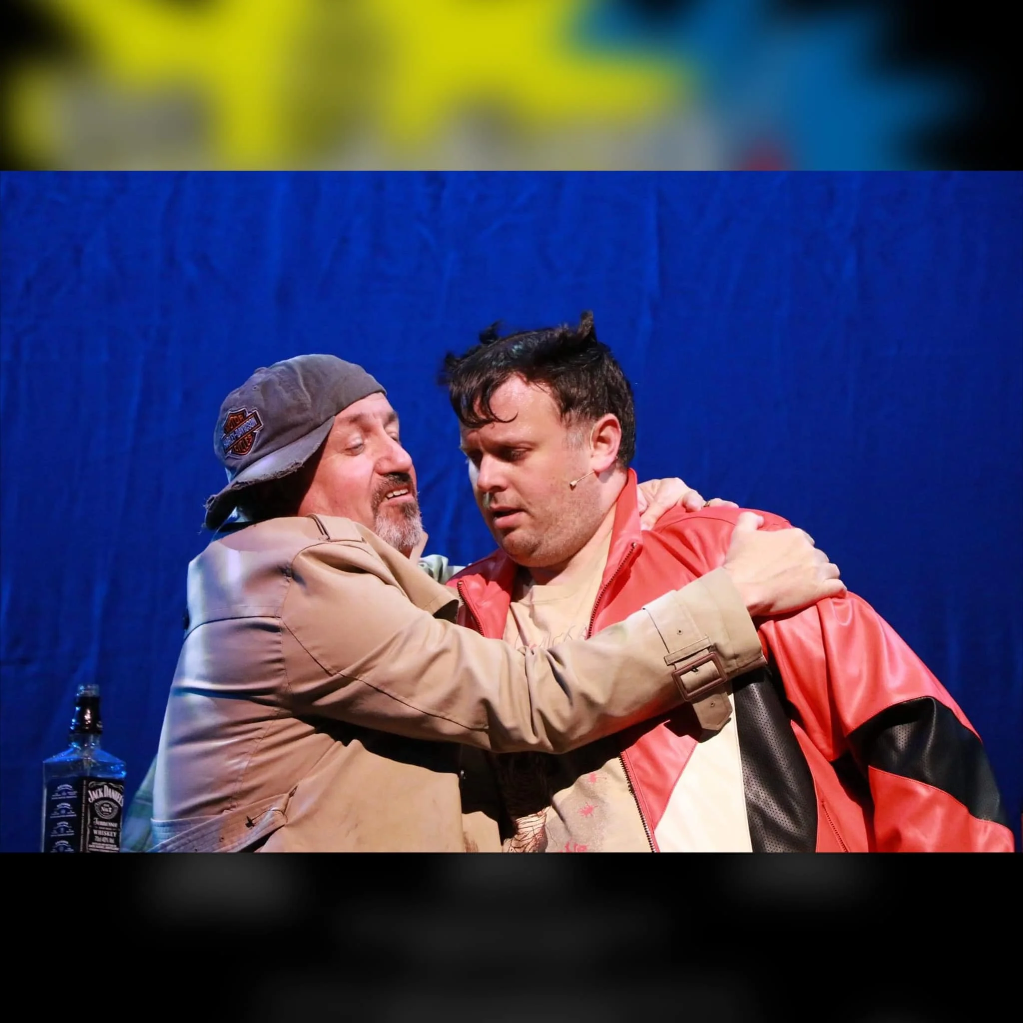 Two people on stage, one women dressed as older man with a beard and gray cap, and the other younger with black hair, in a dramatic scene with blue background