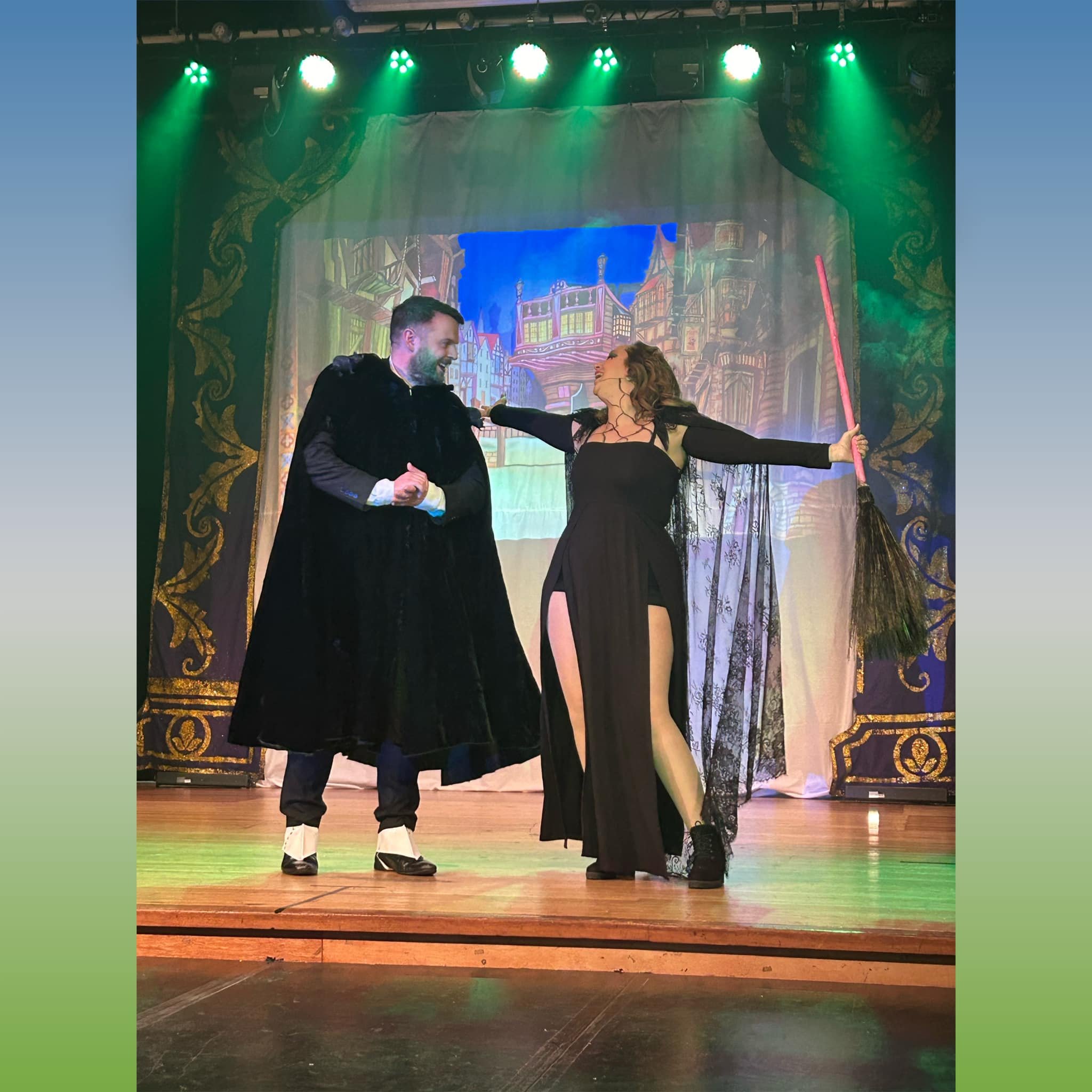 Two actors on stage in costume performing a scene, one man in a black cloak and woman in a black dress with high slit, holding a broom, with a colorful theatrical backdrop.